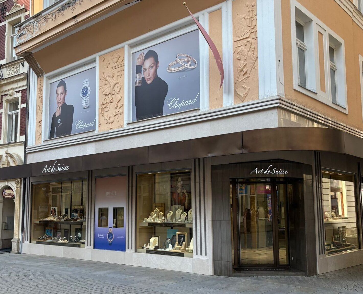alt="Butik Art de Suisse v Karlových Varech s výlohami značek Chopard a Bovet, velkoformátovými reklamami a luxusními šperky a hodinkami"